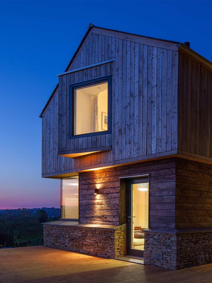 The Larch House exterior at dusk showing larch cladding, stone base and lit interiors, Gloucestershire